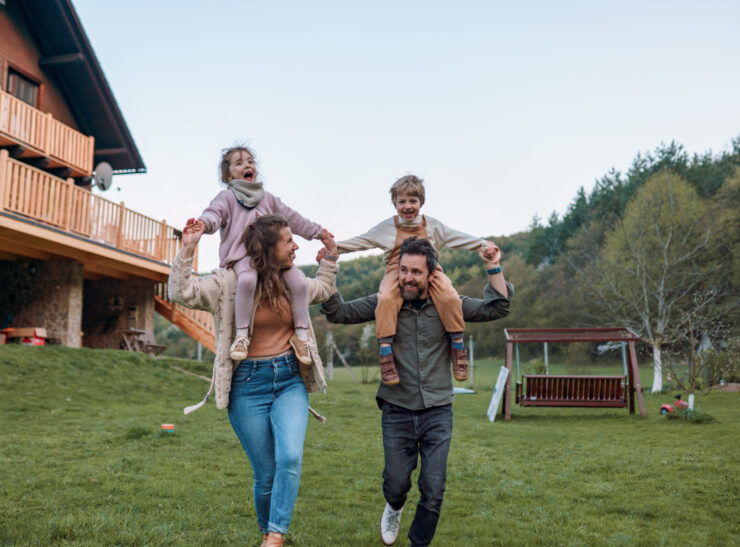 Happy parents with small children on piggybacks running and having fun together in a garden near their house.