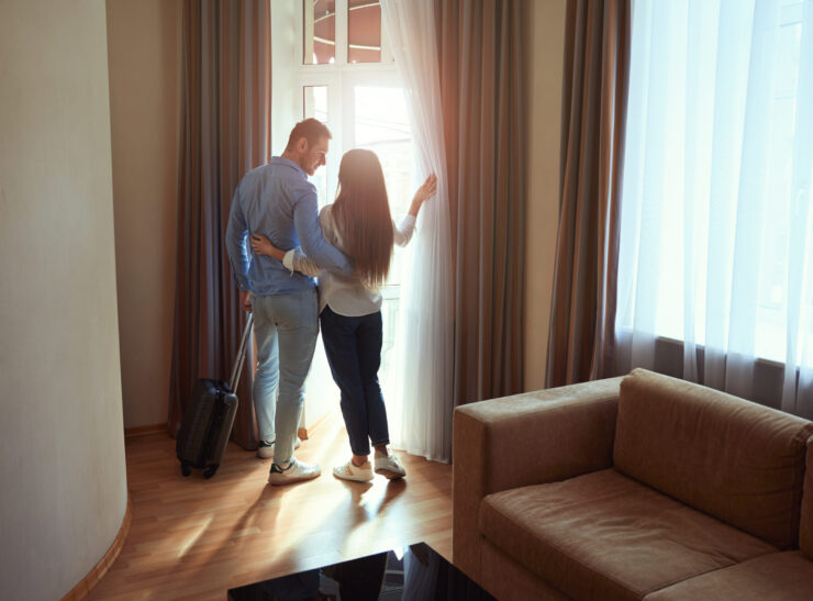 Loving young couple hugging stands with a suitcase in a hotel room and looks out the window